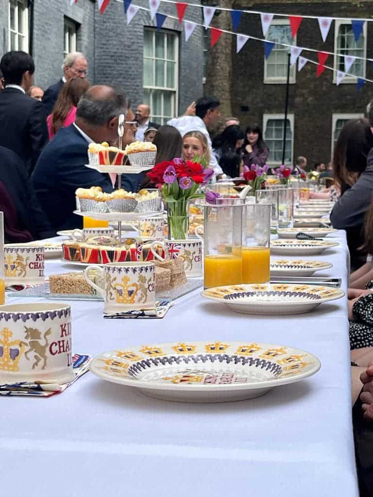 A long white table stretches through a courtyard adorned with red, white and blue bunting. The table is set with decorate 'King Charles III' plates and mugs, trays of cakes, and fresh flowers.