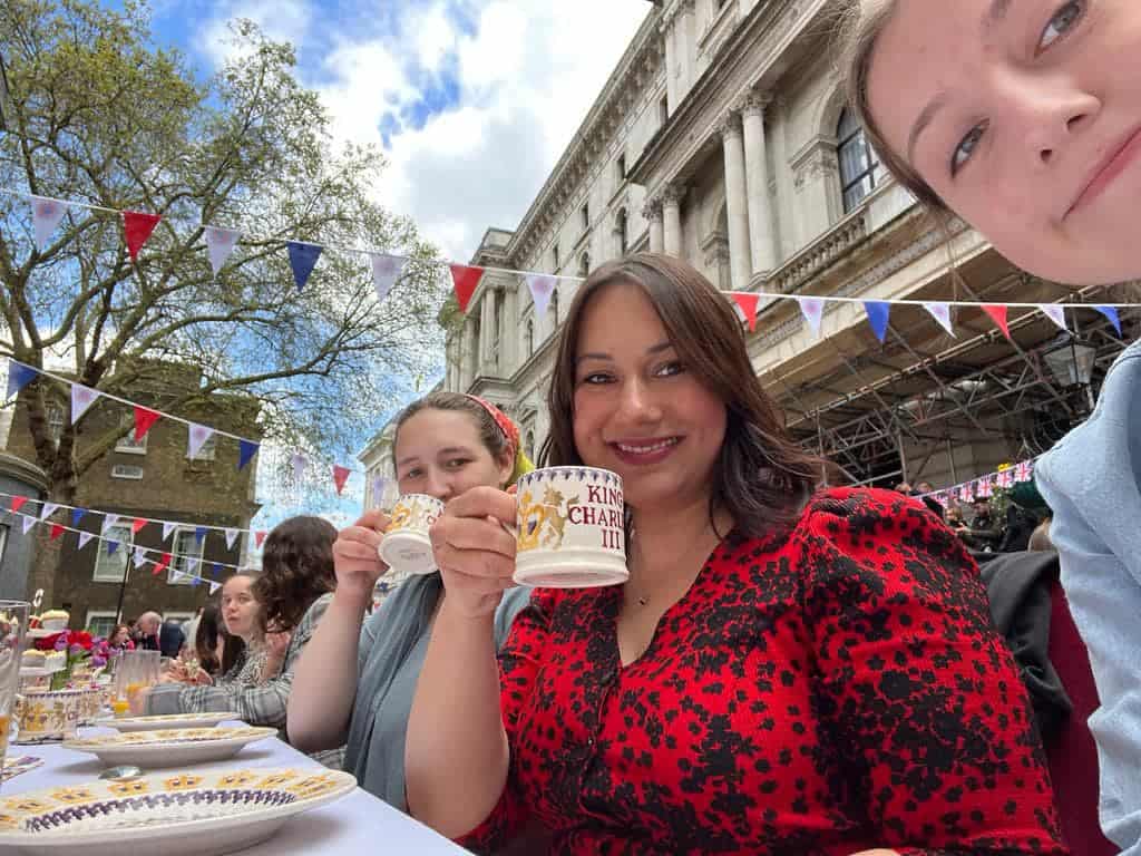 Two smiling young people and a youth worker sip tea from mugs that read, 'King Charles III.' In the backyard is a sunny courtyard adorned with red, white and blue bunting.