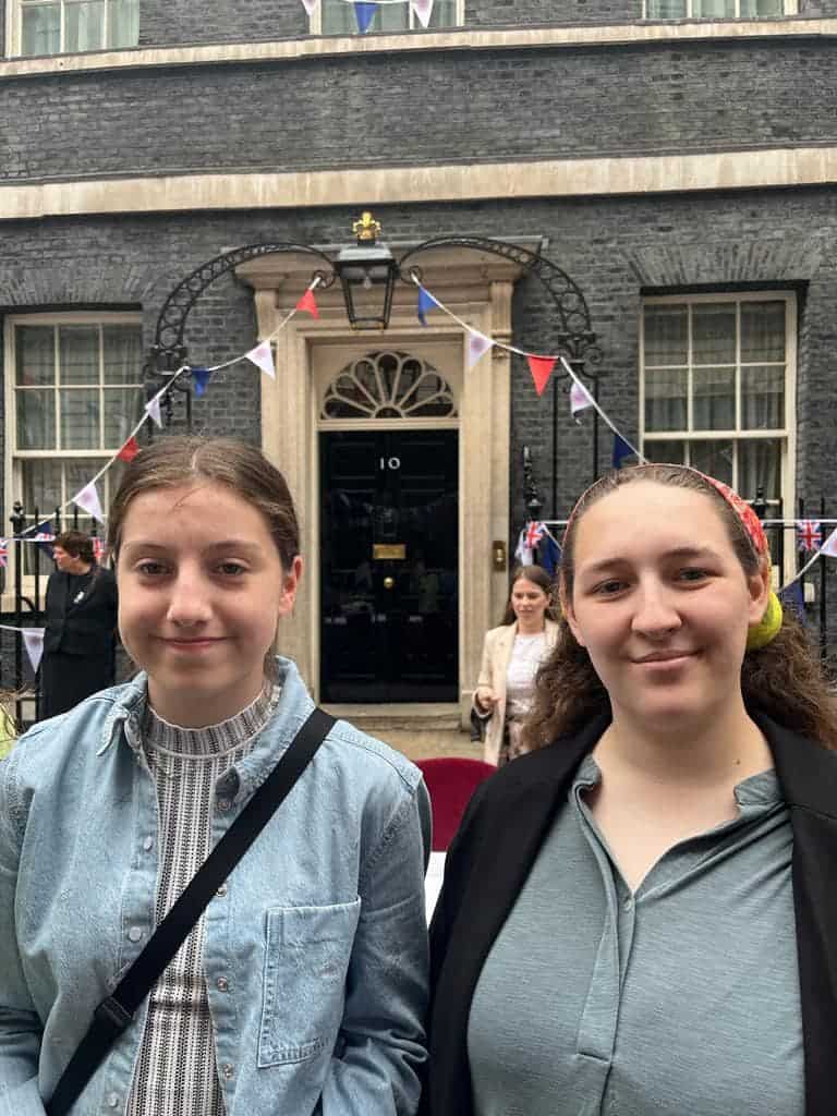 Two young people smile in front of a glossy black door with the number 10 on it. The door has been decorated with red, white and blue bunting.