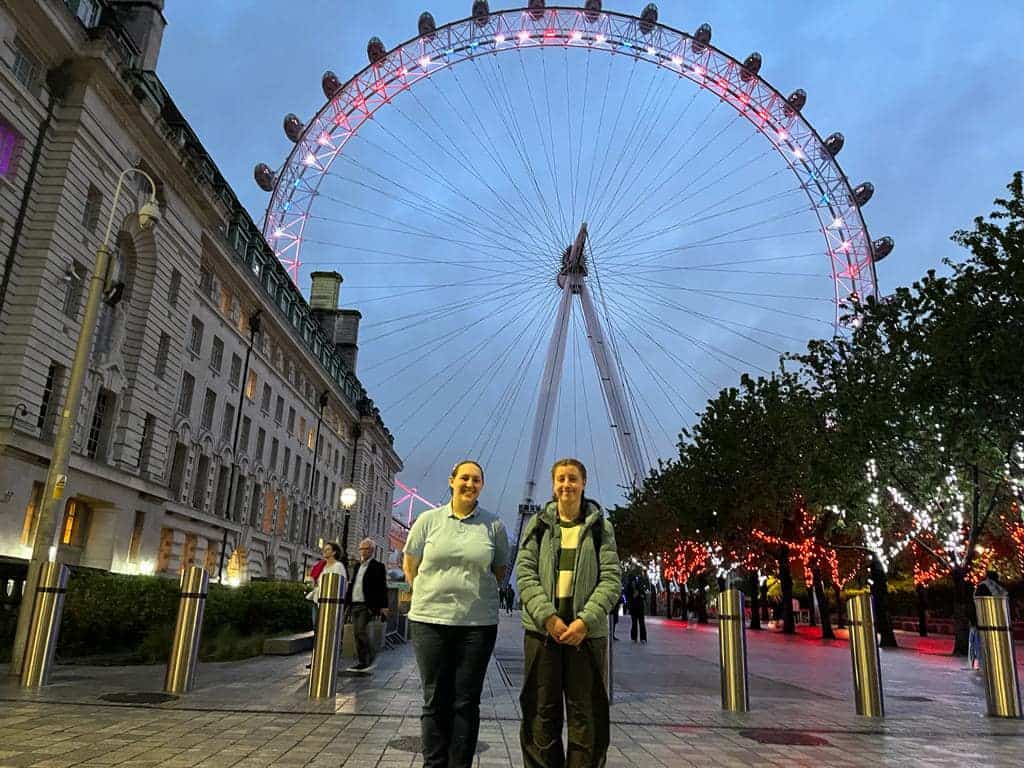 Two young people stand smiling in front of the London Eye Ferris wheel, which has been illuminated red, white and blue. On the right, a line of trees are decorated with white and red lights.