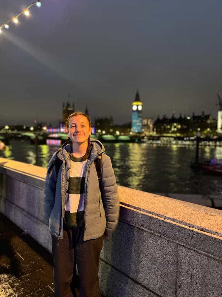 A young person stands smiling next to a low stone wall. In the background is the River Thames and a blurred image of Big Ben and the Palace of Westminster lit with red, white and blue light displays.