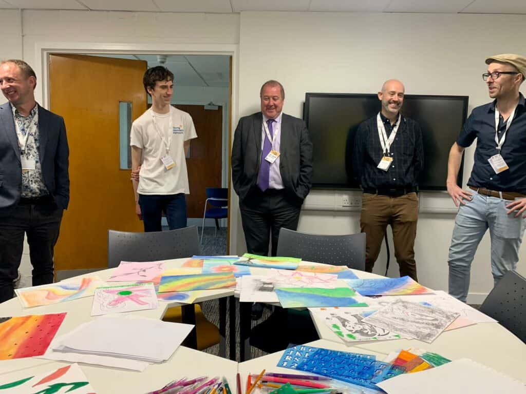 5 people standing in a room with a table in the front full of artwork. people all wearing lanyards and smiling. 