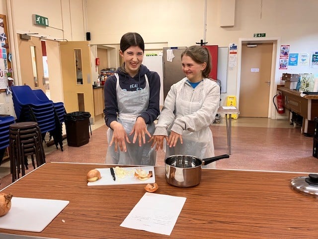 two young people in aprons laughing and pointing open hands downwards.  chopped onion on a chopping board and a silver pan. 