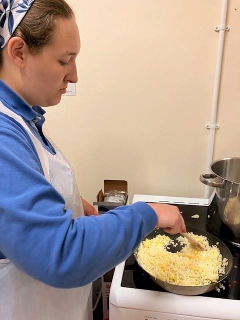 young person in a blue jumper and while apron stirring a pan filled with grated cheese and other ingredients. 