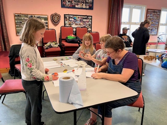 adult helping young people paint rocks at a table. paper, spray bottle and blue roll on table.  people in background, one standing and one sitting.  pink chairs stacked in background. 