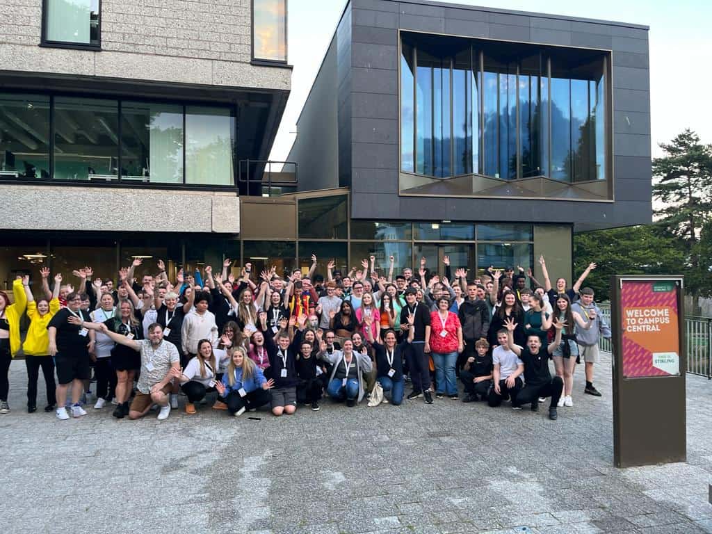 large group of people waving their hands in the air, some kneeling at the from and most standing in a semi-circle shape facing the camera. 