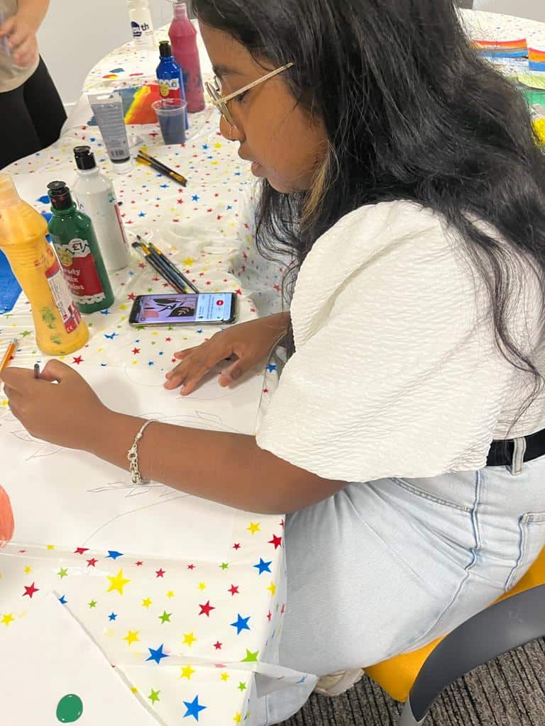 young person sitting at table with paint bottles, drawing a picture from their phone