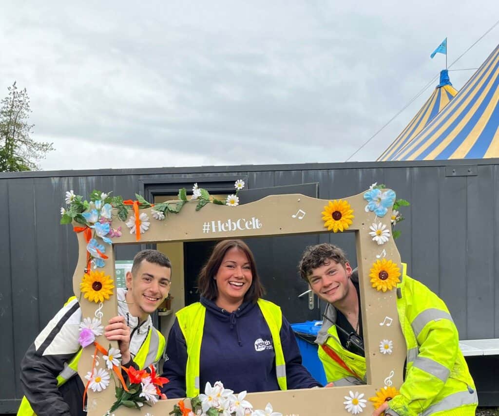 Two young people and a Young Islanders Network employee smile and pose inside a large wooden frame that reads '#HebCelt'