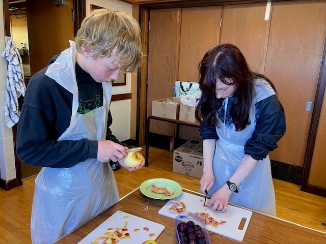 two young people cutting apples and  other food.  wearing white plastic aprons. wooden background, cupboard doors. 
