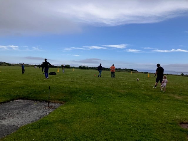 green field, blue skies, adults and children playing football. yellow cones around. 