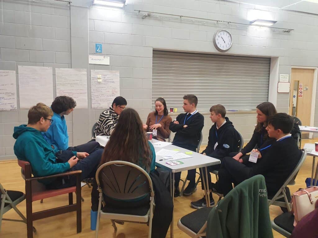 9 people sitting round a table discussing, in a hall with paper on the wall with writing. 