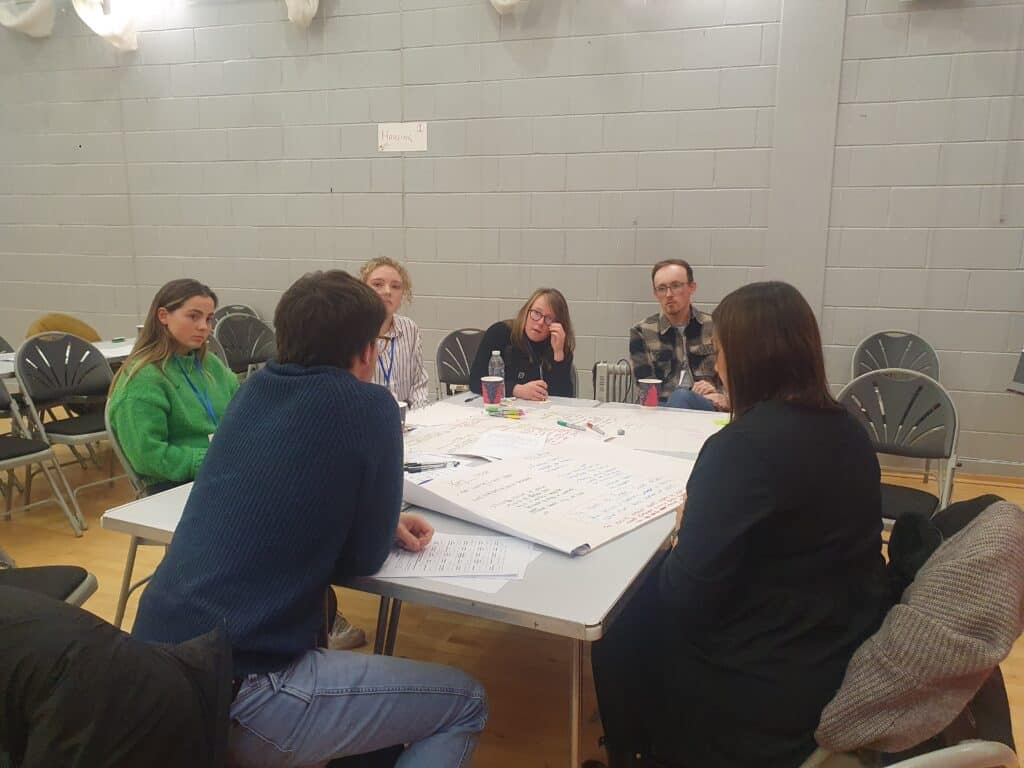 6 people sitting around a table in discussion. one person in centre touching their glassed. table with paper and pens in the middle. white walls in the background