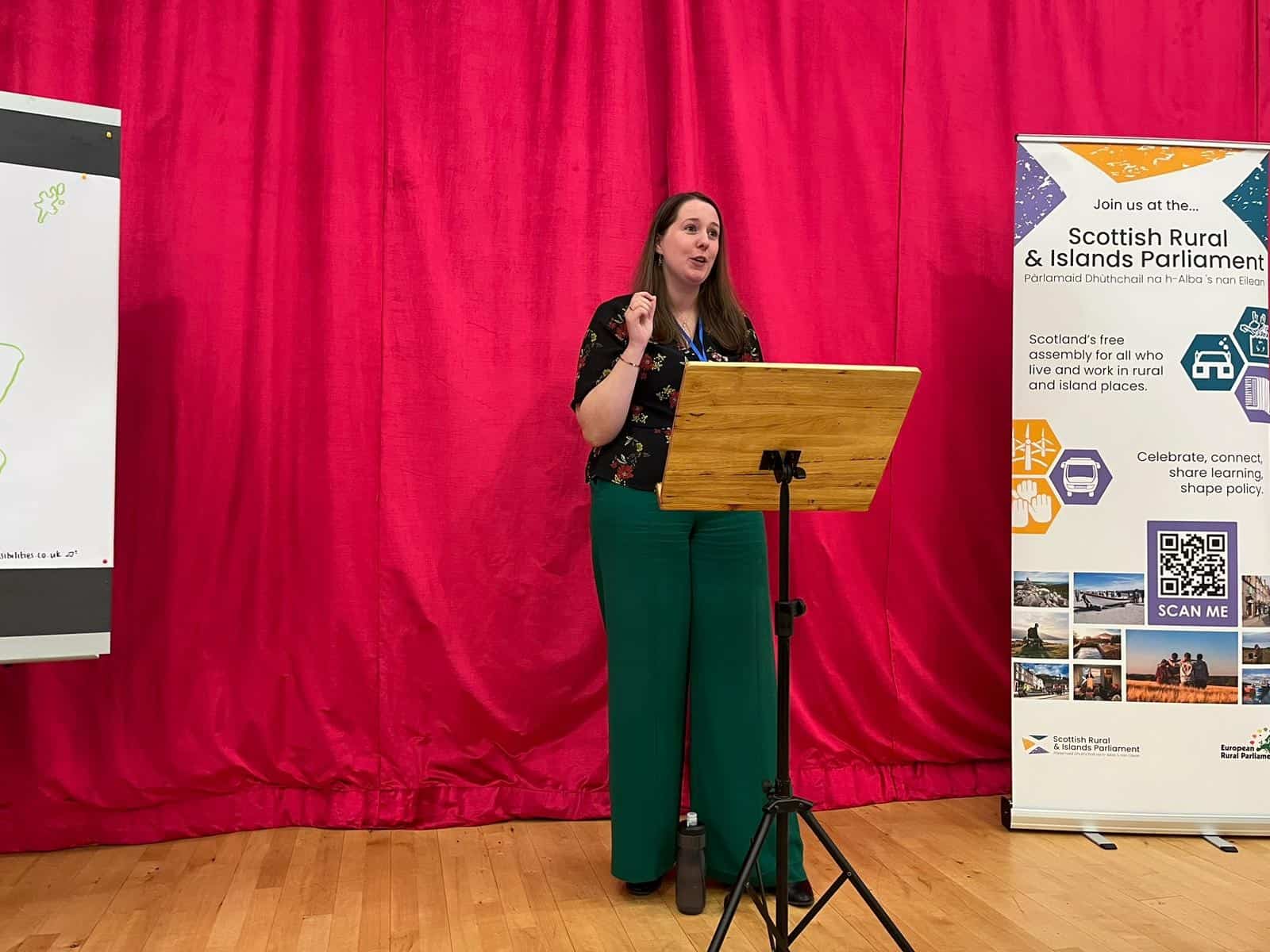 red curtain. female, Emma Roddick,  standing in front of stand delivering a speech.  tall banner for scottish and rural islands parliament to the right of the speaker. 