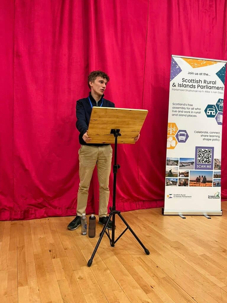 young male holding onto stand delivering a speech in front of a big red curtain. Banner for the Scottish rural and islands parliament to the right. 
