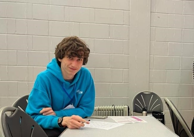 young person in a blue hoodie smiling at the camera while sitting at a table alone. holding a pen and a piece of paper on the table. white brick wall behind him. 