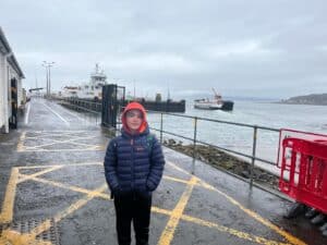 A boy standing hands in pocket, hood up on pier with two Calmac ferries in background.