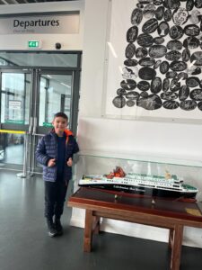 Departures written on the wall over a door. A boy standing by a model of a ship in a case. 