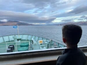 A boy looking out of the window over the front of a ship with islands in the distance and a lighthouse in daylight.