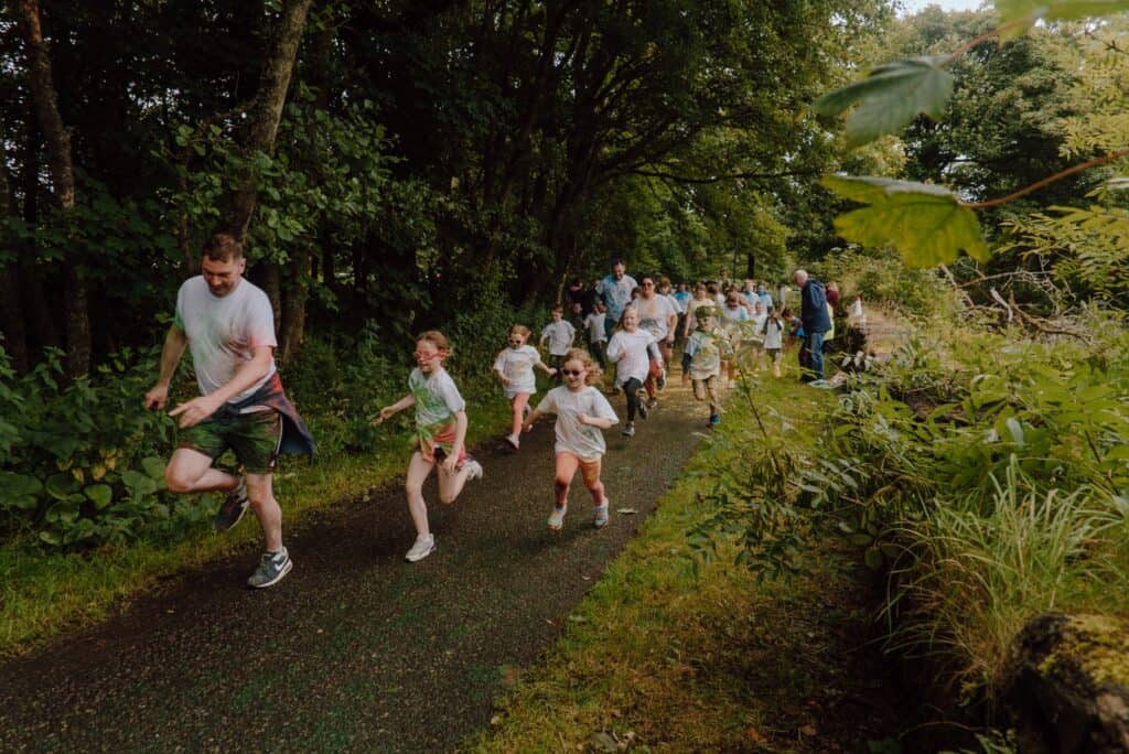 A large group of people from young children to adults run through the woods on a colour dash, covered in a rainbow of coloured dust.