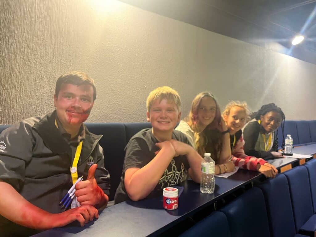 five young people smiling at camera while sitting at the back of a lecture theatre. one young person with alot of special effects make up